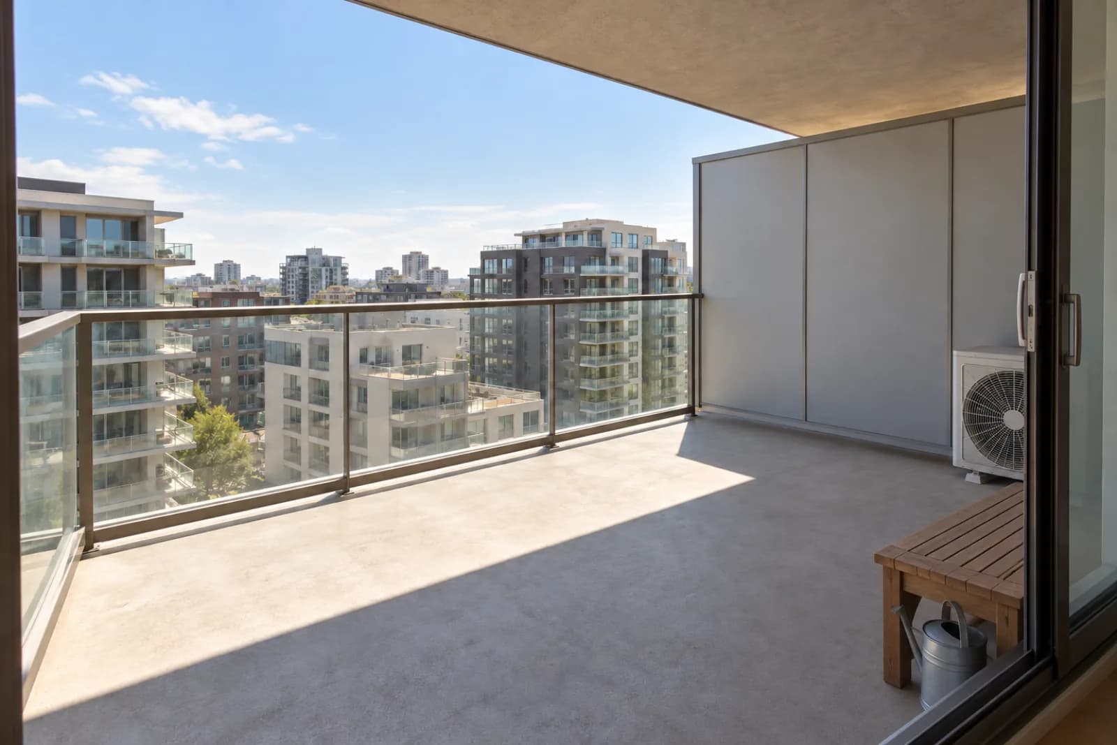 An empty fourth-floor apartment balcony on a summer afternoon, seen from the open balcony door: a concrete floor, a glass railing with a metal top rail, a solid privacy screen at the right-hand end with an air-conditioner condenser in that corner, and the balcony above shading the back half of the floor.