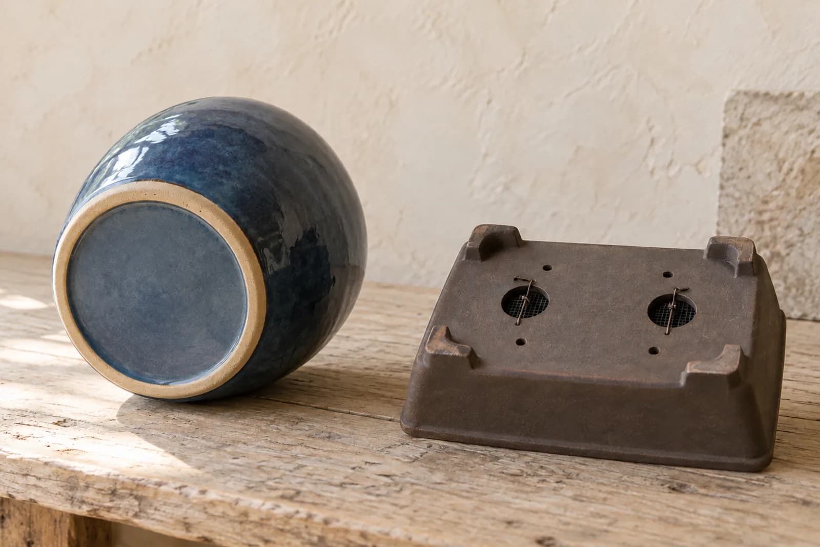 Two empty pots on a wooden potting bench. On the left, a glossy glazed decorative pot lies on its side with its solid base, which has no hole, turned toward the camera. On the right, a shallow brown bonsai pot turned upside down shows two large drainage holes with plastic mesh showing through them, small wire holes, and four short feet.